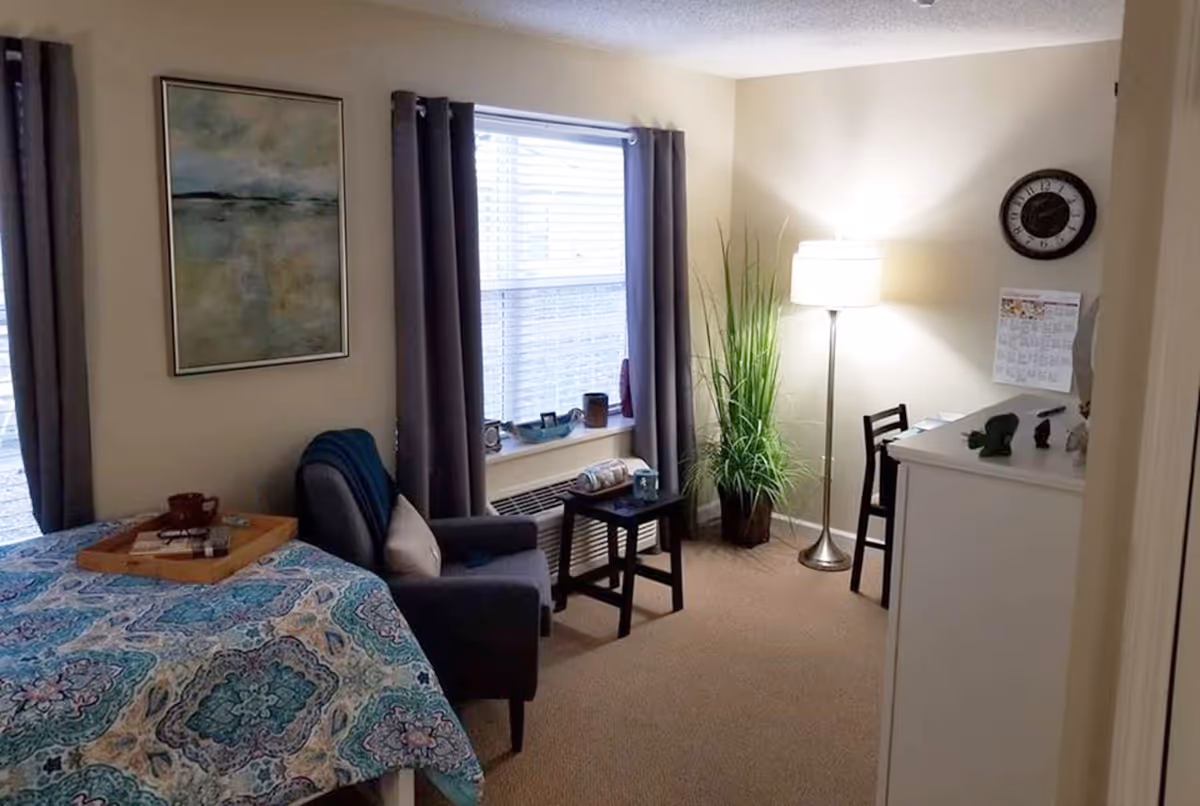 A cozy bedroom with a bed covered in a colorful patterned bedspread, a gray armchair with a pillow, a small black side table, a window with gray curtains, a tall floor lamp, a potted plant, a white dresser with decorative items, a wall clock, and a calendar on the wall.