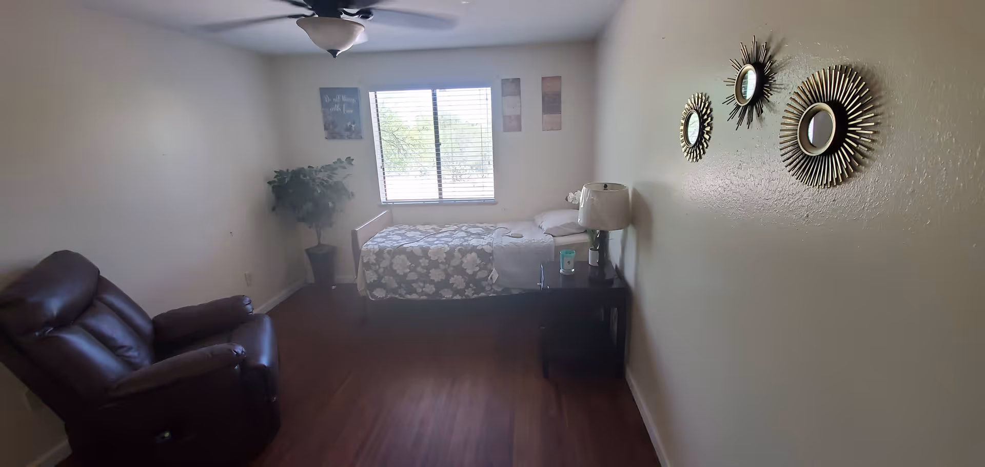 A small, simple bedroom with a single bed covered in a floral bedspread positioned against the far wall under a window with blinds. To the right of the bed is a dark wooden nightstand with a lamp and some items on it. On the left side of the room is a dark brown recliner chair. The walls are light-colored with three decorative sunburst mirrors hanging on the right wall and framed artwork on the wall near the window. The floor is wooden.
