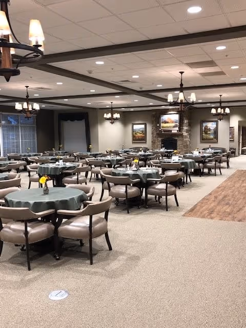 A spacious dining room with multiple round tables covered with green tablecloths, each surrounded by cushioned chairs. Small flower vases are placed on the tables. The room features chandeliers hanging from the ceiling, a stone fireplace with framed paintings above it, and large windows with curtains.