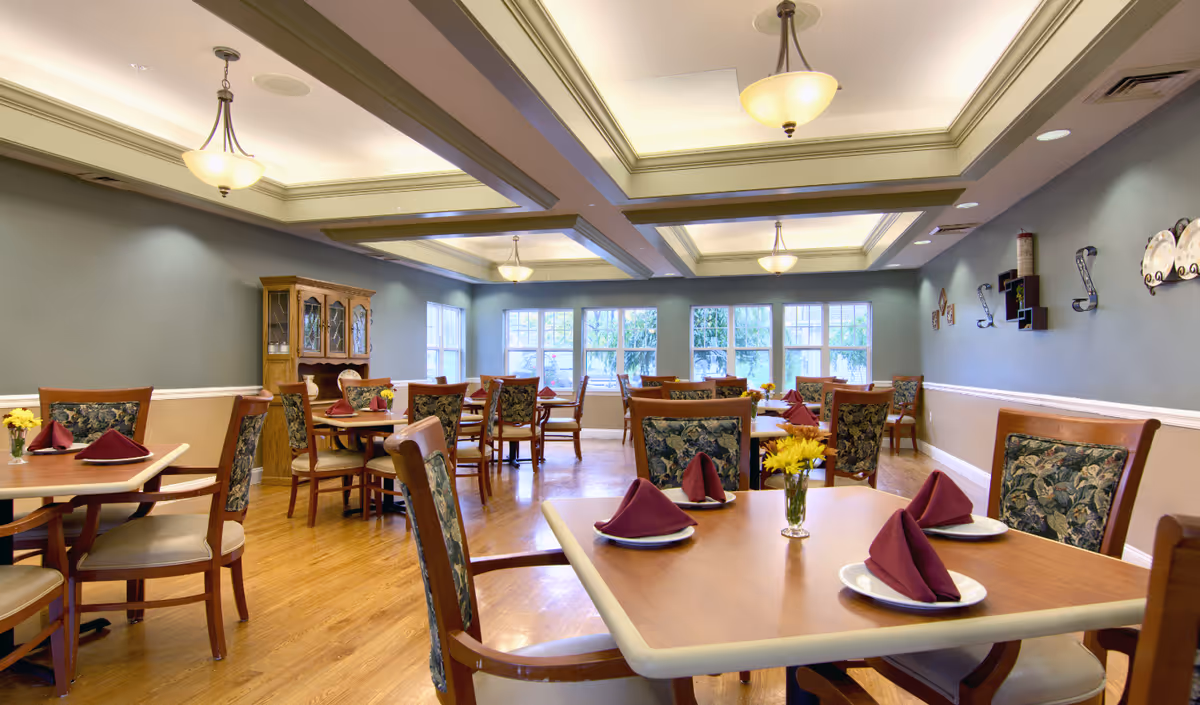 A well-lit dining room with multiple wooden tables and chairs arranged neatly. Each table is set with white plates and folded burgundy napkins. There are small vases with yellow flowers on the tables. The room has large windows letting in natural light, a wooden floor, and soft blue-gray walls with decorative wall hangings. Ceiling lights provide additional illumination.