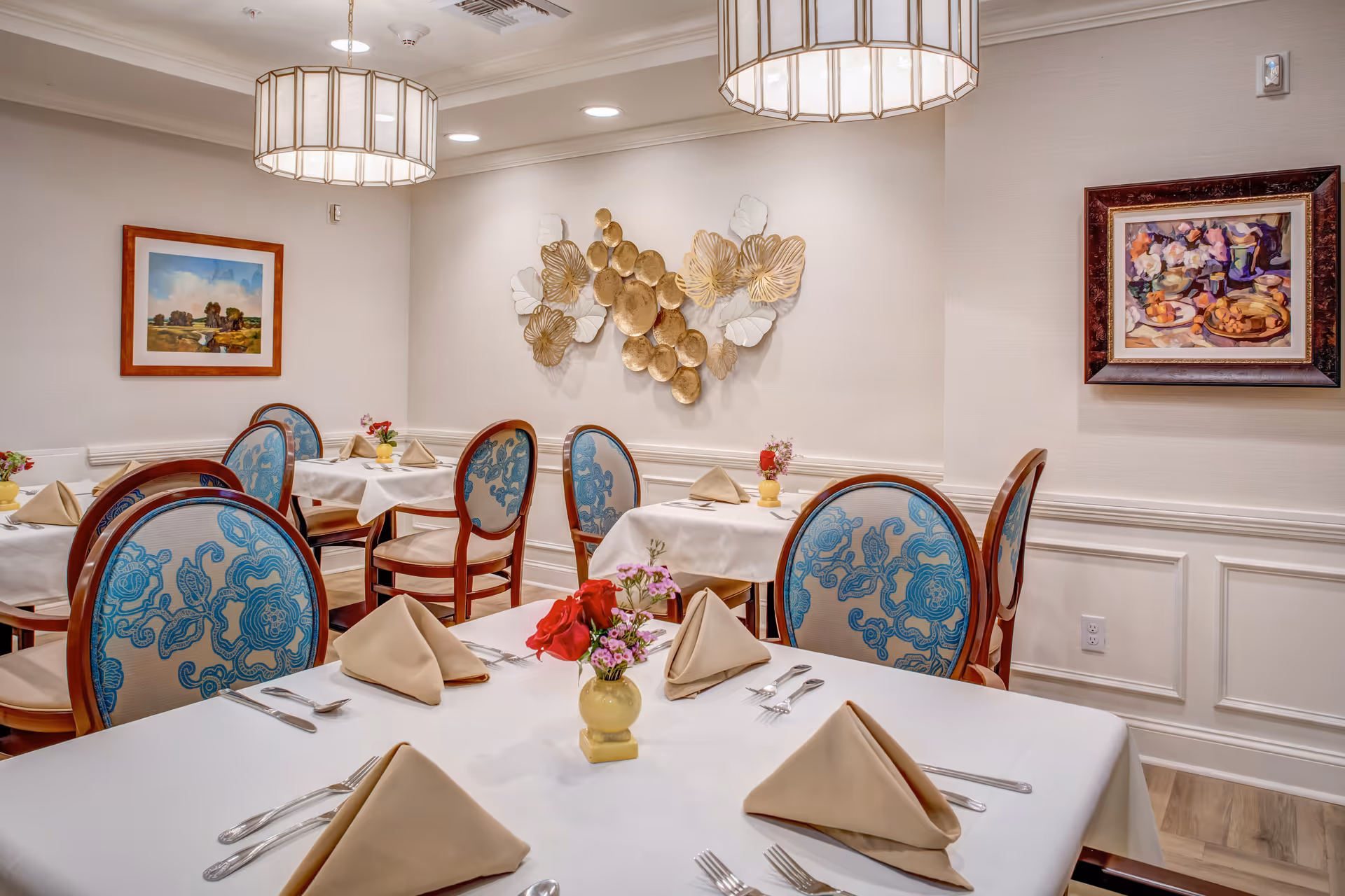 Elegant dining room with round-backed chairs, white tablecloths, folded napkins, floral centerpieces, and wall art.