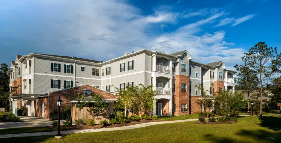 Three-story brick-and-siding retirement community building with balconies, landscaped lawn, and a walkway under a blue sky.