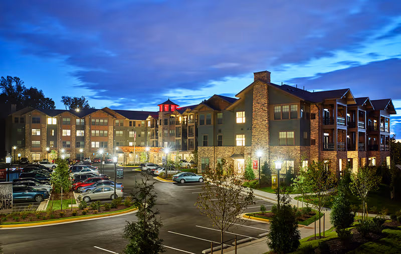 Exterior view of a large senior living facility building at dusk with multiple floors, lit windows, a parking lot with cars, landscaped greenery, and a partly cloudy sky.