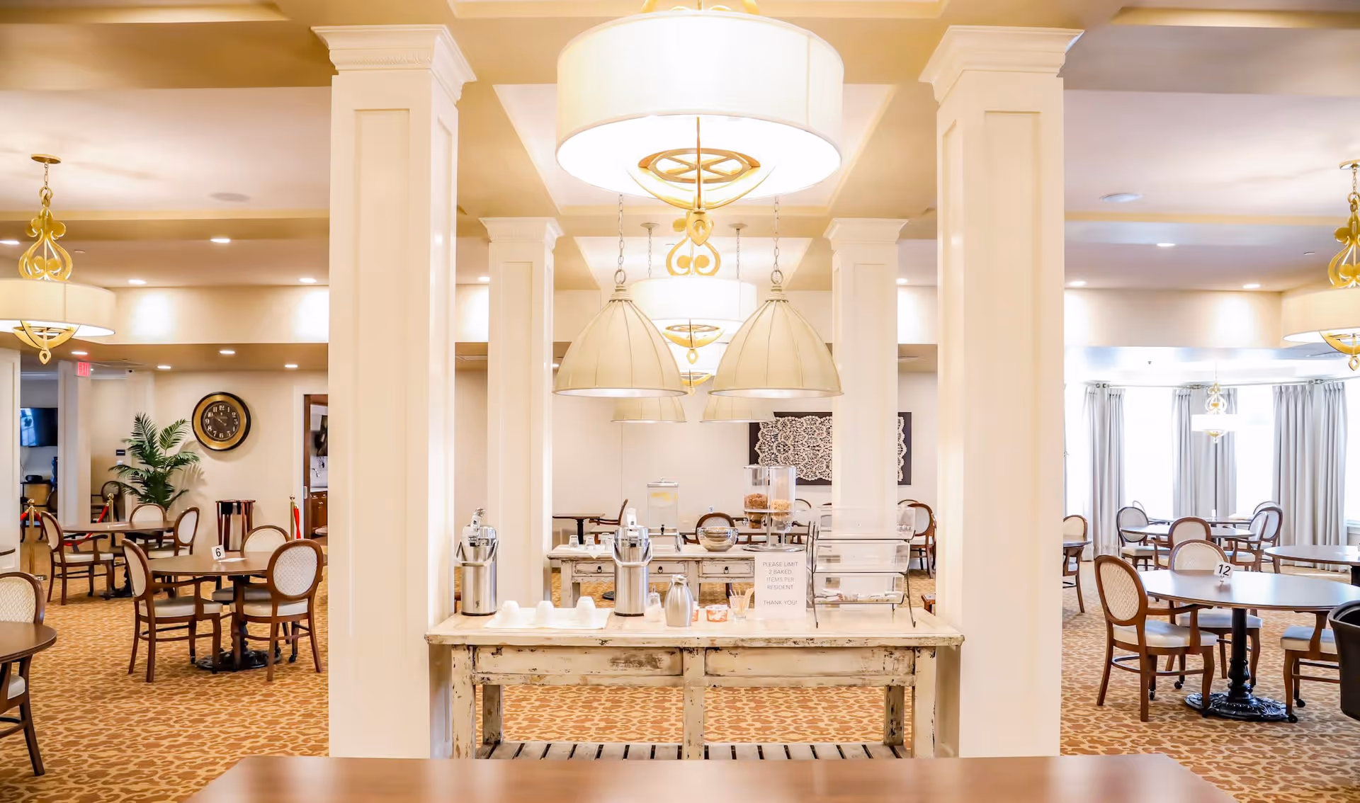 A bright and spacious dining room in a senior living facility with multiple round tables and chairs arranged neatly. The room features beige walls, patterned carpet, large hanging light fixtures, and a central buffet table with dispensers and cups. There are decorative columns and framed artwork on the walls, with natural light coming through windows covered by curtains.
