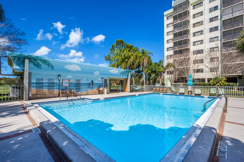 Outdoor swimming pool with clear blue water at a senior living facility. The pool area is surrounded by a concrete deck with lounge chairs and a building with a mural of a beach scene. Palm trees and a tall residential building are visible in the background under a bright blue sky with scattered clouds.