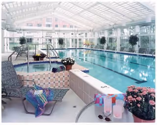 Indoor swimming pool area with clear water, surrounded by large windows and a glass ceiling allowing natural light. There are lounge chairs with towels and bags, potted plants, and a hot tub adjacent to the pool.
