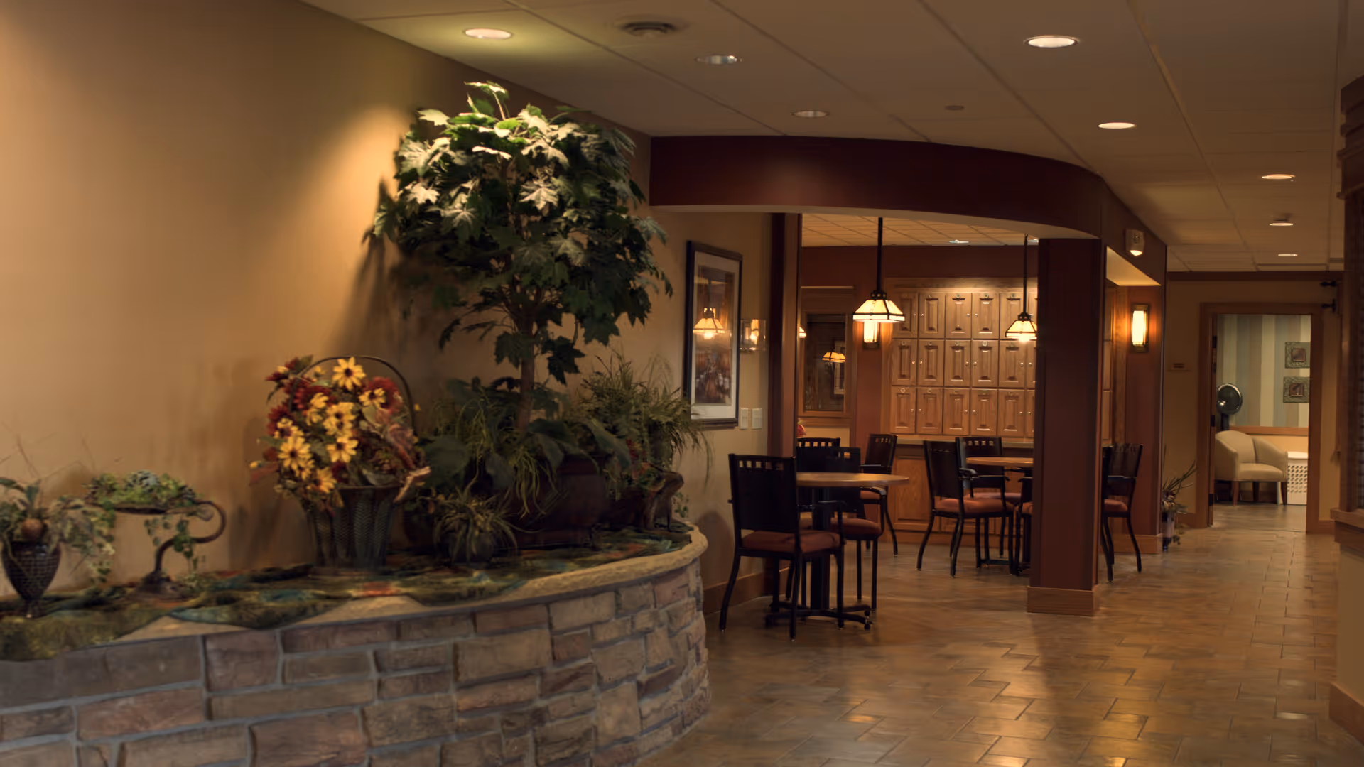 Interior view of a senior living facility hallway with a stone planter filled with various plants and flowers on the left. The hallway leads to a dining area with tables and chairs, warm lighting, and wooden cabinetry in the background. The walls are painted in neutral tones, and the floor is tiled.