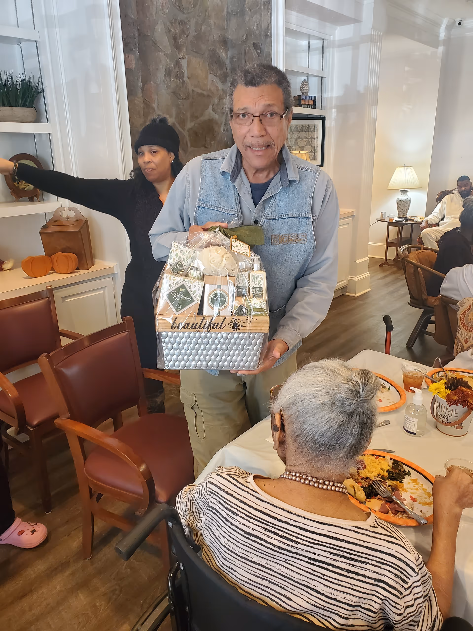 A man holds a wrapped gift basket while residents sit at a dining table in a senior living common room.