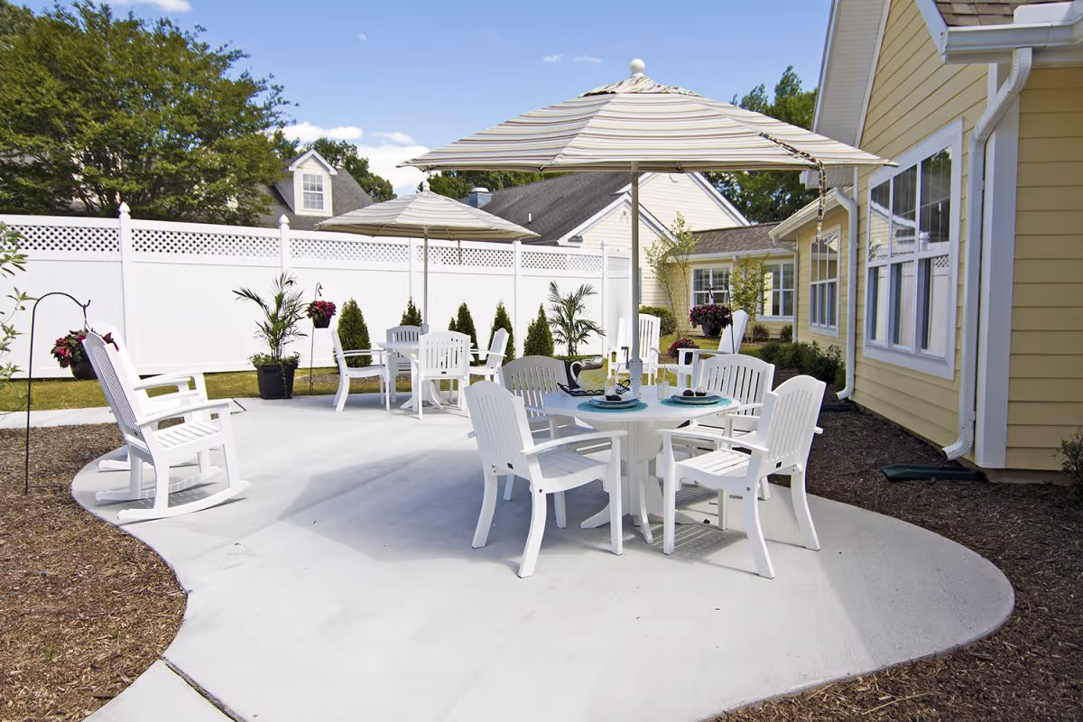 Outdoor patio area with white plastic chairs and tables under striped umbrellas, surrounded by a white fence and landscaping with potted plants and bushes, adjacent to a yellow building.