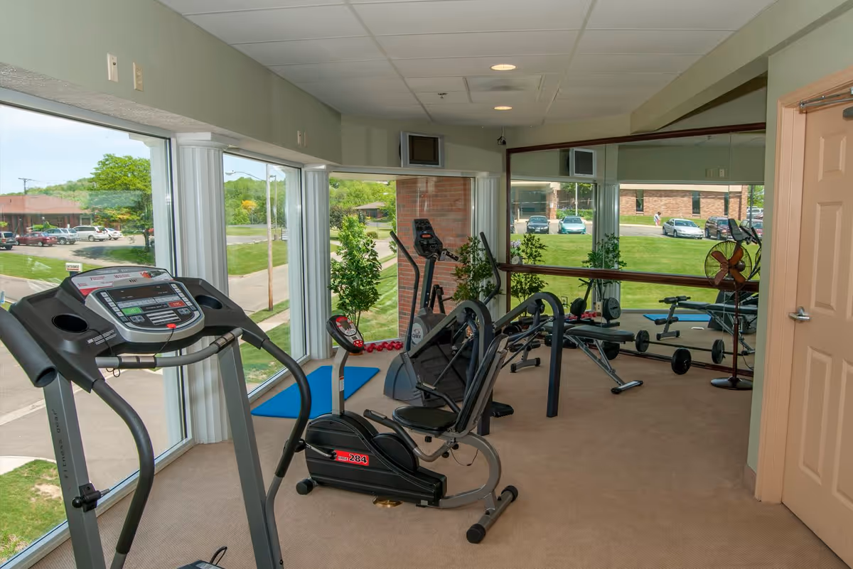 A small fitness room with exercise equipment including a treadmill, stationary bike, weight bench with dumbbells, and an elliptical machine. The room has large windows providing a view of the outside parking lot and greenery. There is a large mirror on one wall and a closed door on the right side.
