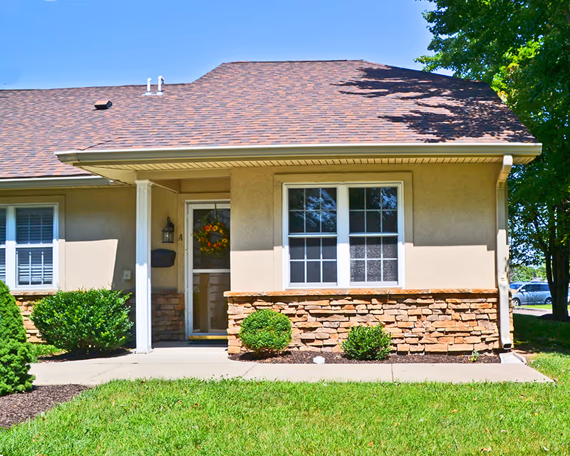 Exterior view of a single-story residential building with beige walls and a brown shingled roof. The entrance door has a decorative wreath, and there are two windows with white frames. Small bushes and green grass are in front of the building, with a concrete walkway leading to the door. Trees and a parked car are visible in the background.