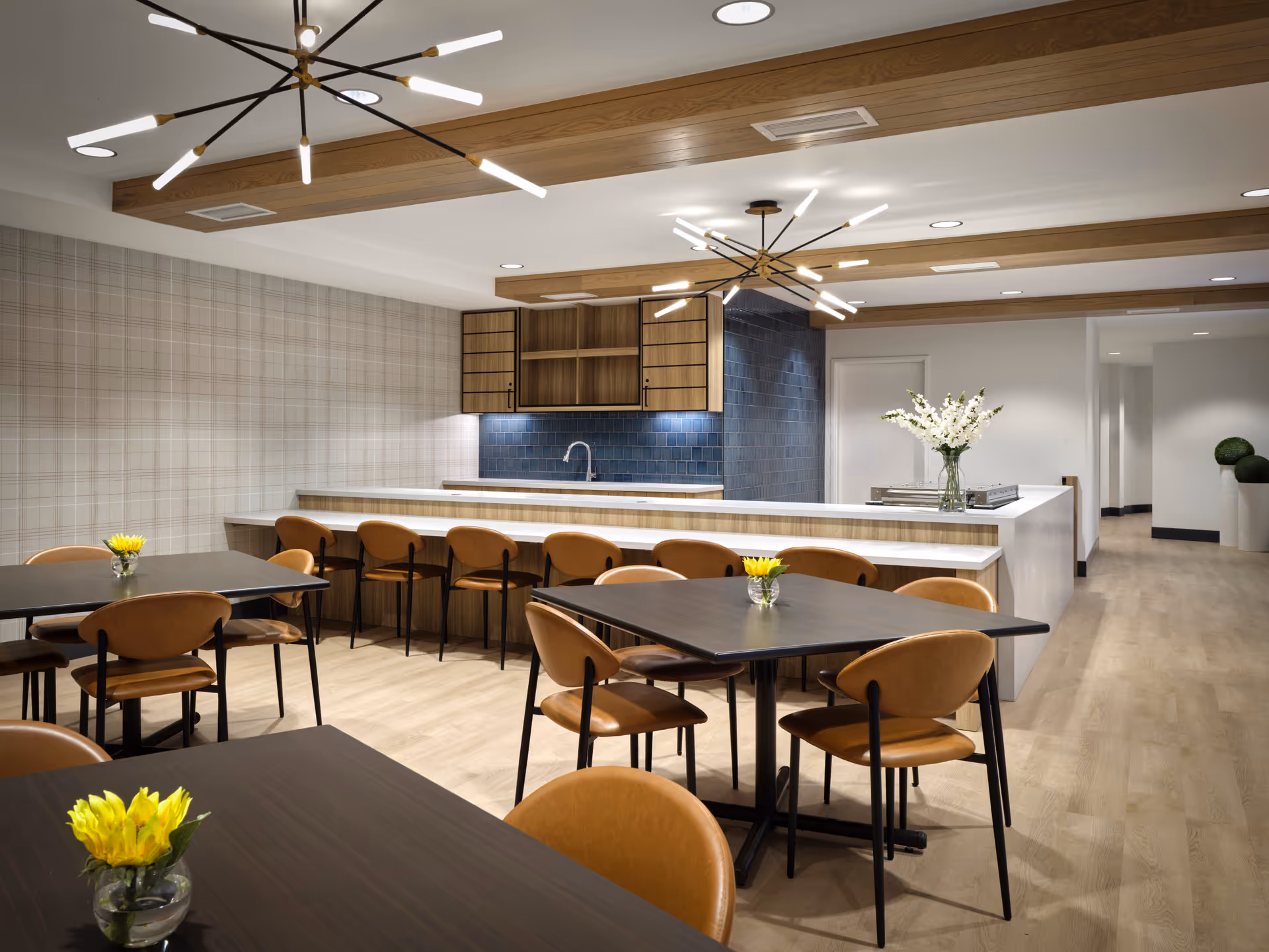 A modern dining area with several dark wooden tables and brown leather chairs. Each table has a small vase with yellow flowers. The room features a long counter with bar stools, a kitchen area with open wooden cabinets, blue tiled backsplash, and two contemporary ceiling light fixtures. The floor is light wood, and the walls are decorated with a subtle plaid pattern and white paint.
