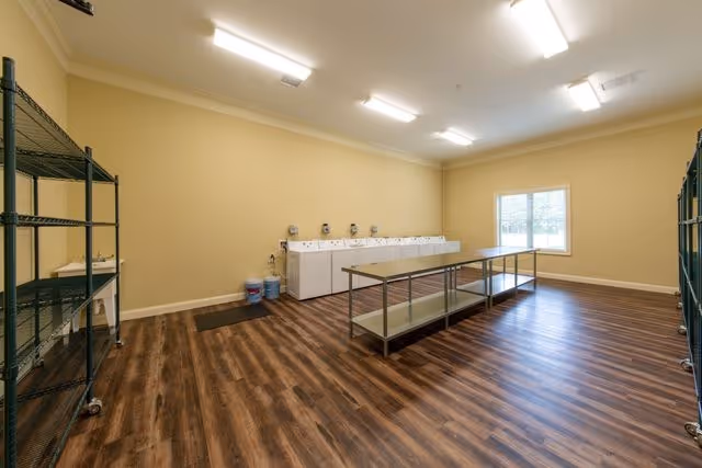 Spacious laundry/utility room with a row of washing machines along the back wall, central folding tables, metal shelving, and a window.