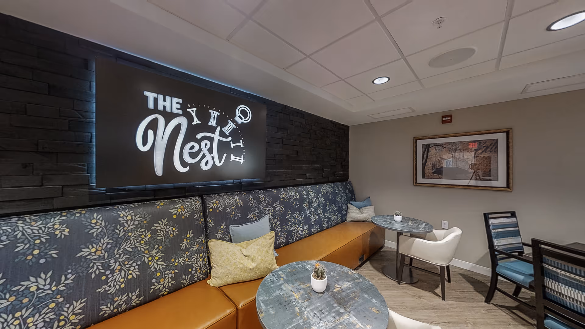 A cozy seating area in a senior living facility featuring a long cushioned bench with floral patterned upholstery and several throw pillows. Two small round tables with potted plants are placed in front of the bench, accompanied by a white armchair and a wooden chair with blue cushions. A framed picture hangs on the beige wall, and a backlit sign on a dark stone wall reads 'The Nest' with clock elements.