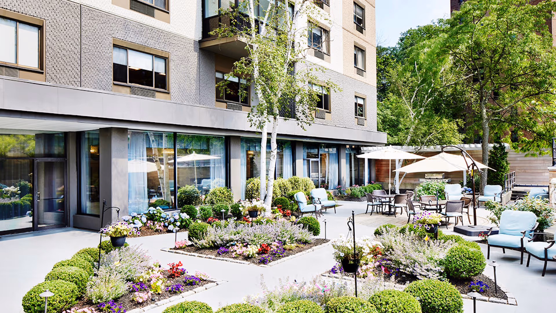Outdoor courtyard area at Atria Stamford featuring well-maintained flower beds with colorful flowers and green shrubs, several seating areas with cushioned chairs, tables with umbrellas, and trees providing shade, adjacent to a multi-story building with large windows.