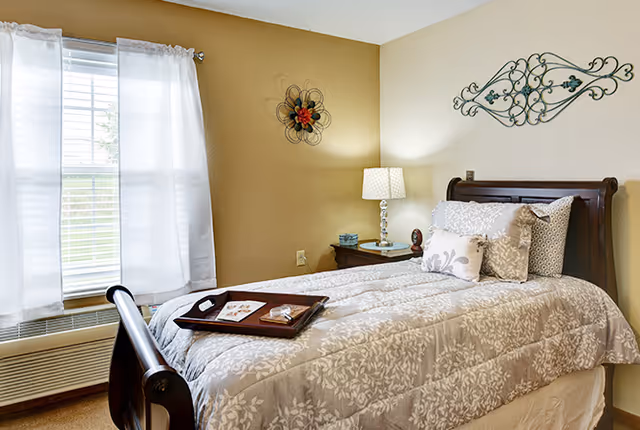 A cozy bedroom with a single bed featuring a floral patterned comforter and multiple pillows. A wooden nightstand beside the bed holds a lamp and a small clock. The room has light-colored walls decorated with metal wall art, and a window with white curtains allowing natural light to enter.