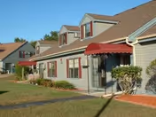 Exterior view of a single-story residential building with gray siding, red window shutters, and red awnings over the entrances. The building is surrounded by a well-maintained lawn and some shrubs under a clear blue sky.