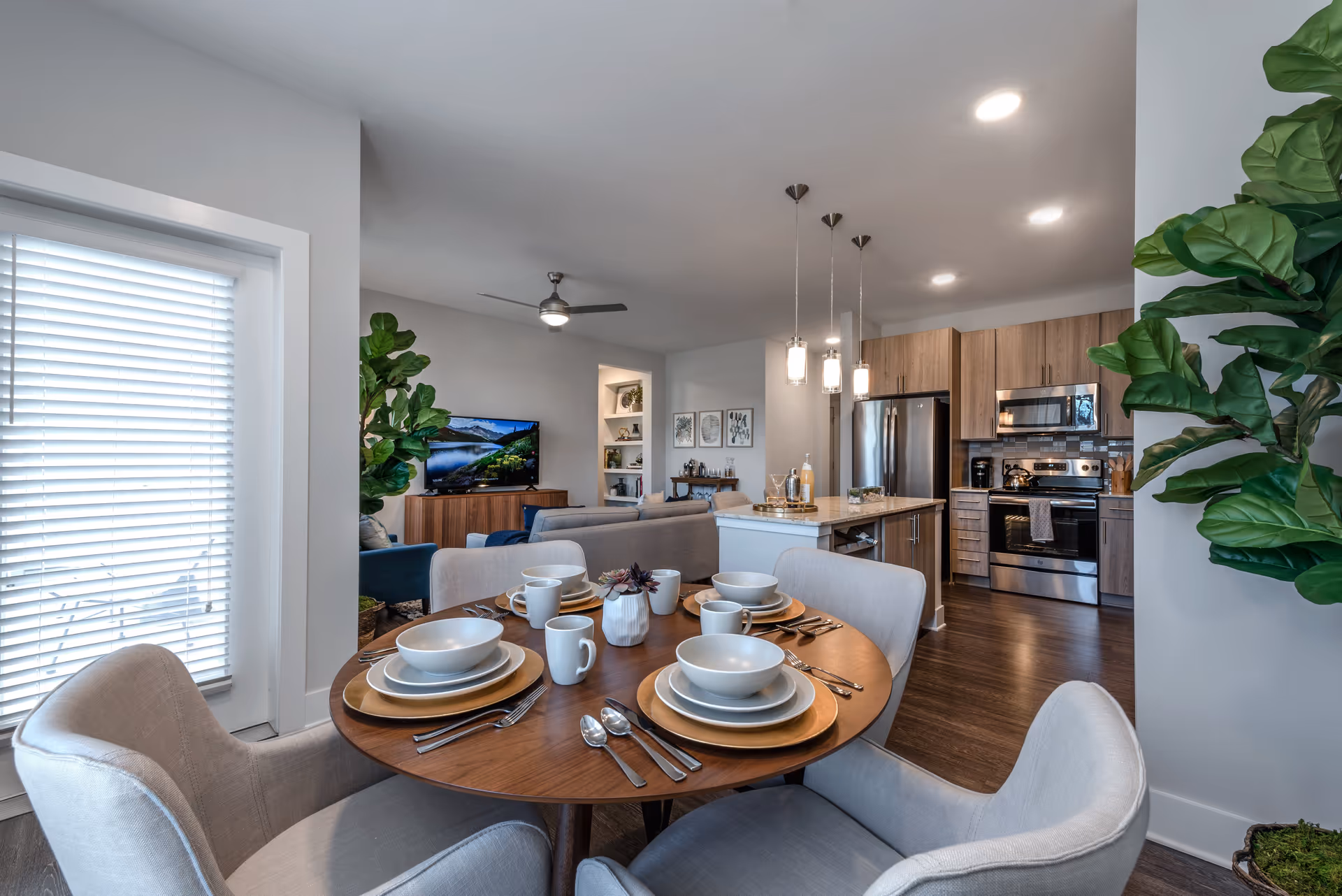 Open-concept apartment interior showing a round dining table set for four in the foreground with a living area, kitchen island, and stainless steel appliances in the background.
