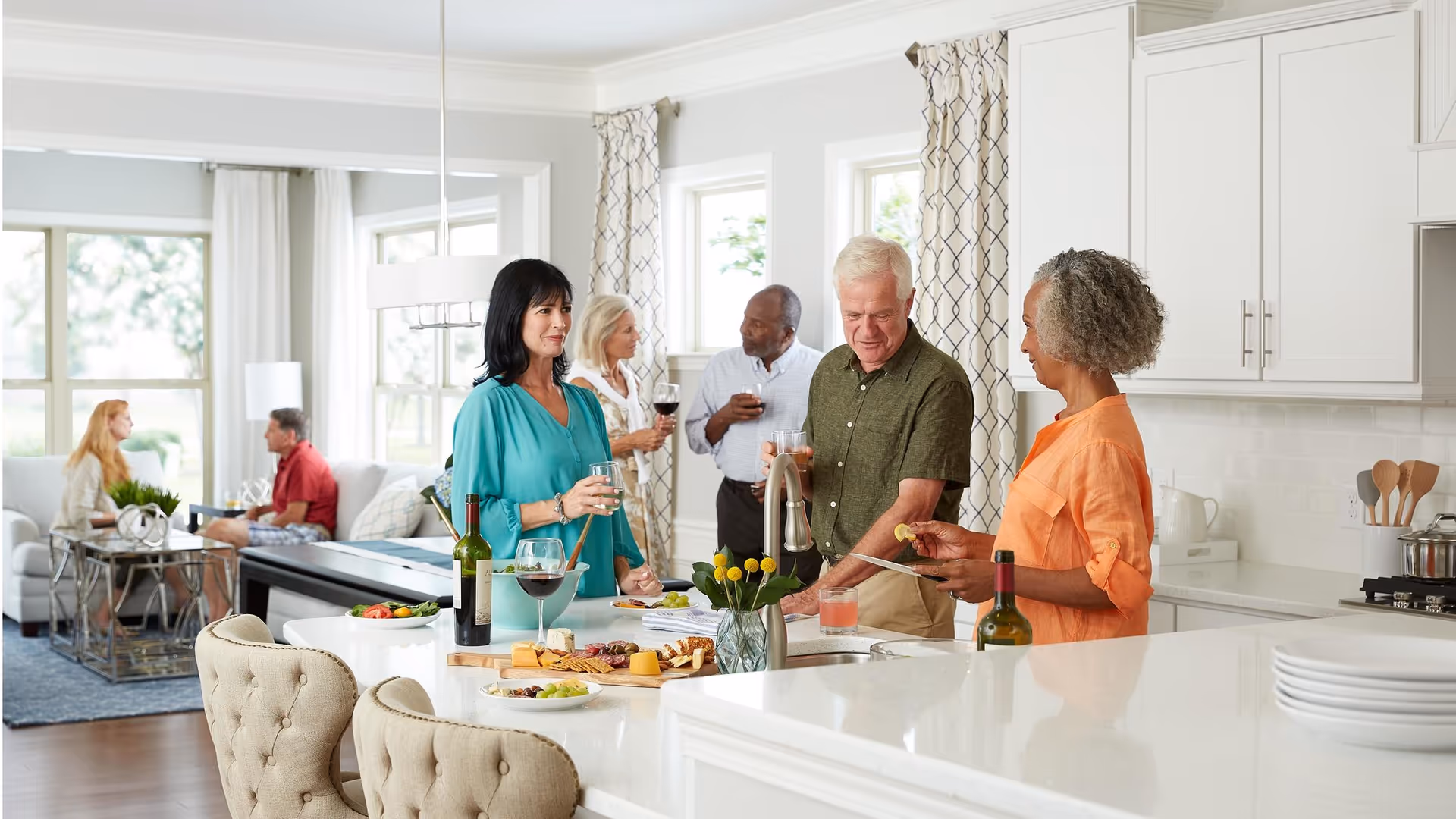 A group of older adults socializing in a bright, modern kitchen and living room area. Three people stand around a kitchen island with drinks and snacks, while two others sit on a couch in the background engaged in conversation. The space features white cabinetry, large windows with patterned curtains, and a clean, inviting atmosphere.
