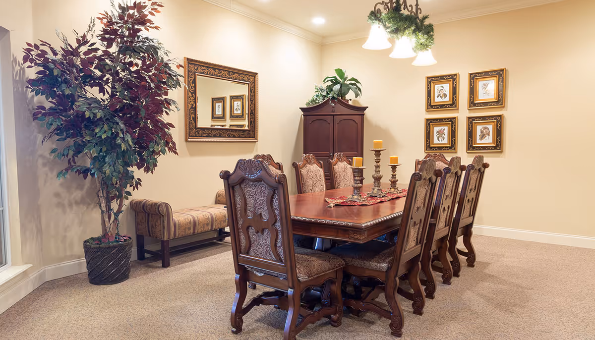 A formal dining room with a large wooden dining table surrounded by eight ornate upholstered chairs. The table is decorated with a centerpiece featuring candles on candle holders. On the wall, there is a large decorative mirror and four framed botanical prints. A wooden cabinet with a plant on top is placed against the wall. A large potted plant is positioned near a window, and a cushioned bench is placed under the mirror. The room is softly lit by ceiling lights and a hanging light fixture with greenery.