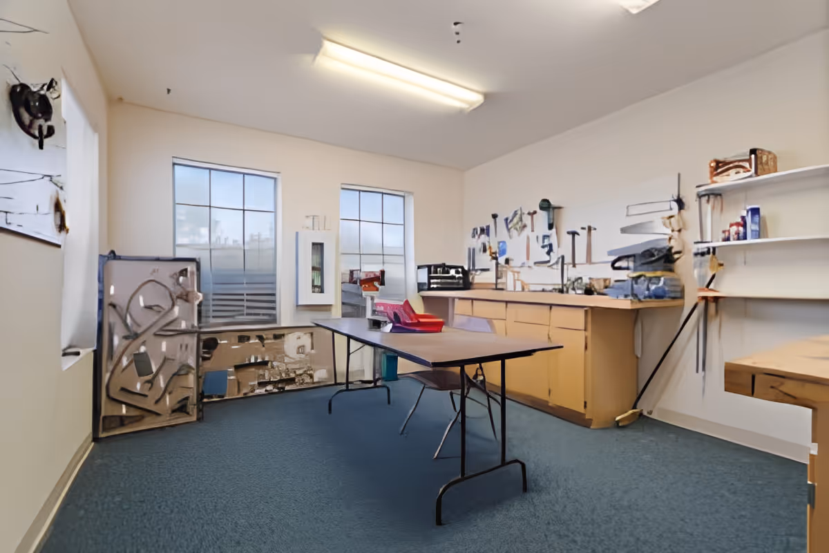A workshop room with a long folding table and chairs in the center, a countertop with cabinets underneath, and various tools hanging on the wall. The room has two large windows letting in natural light and shelves with supplies on the right side.
