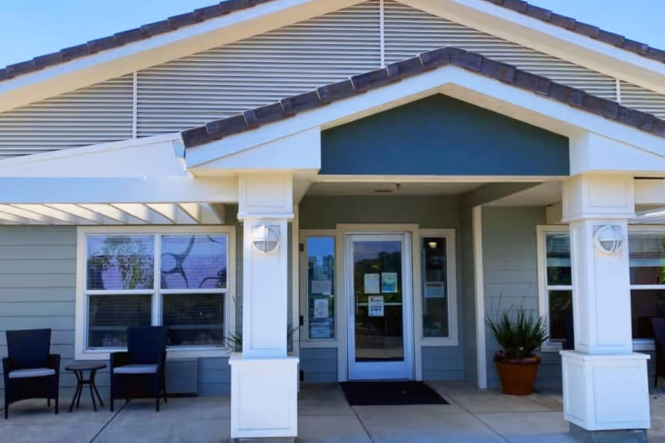 Front entrance of Lassen House Senior Living facility showing a covered porch with two white pillars, a glass door with notices posted, two black chairs with a small table on the left side, and a potted plant on the right side.