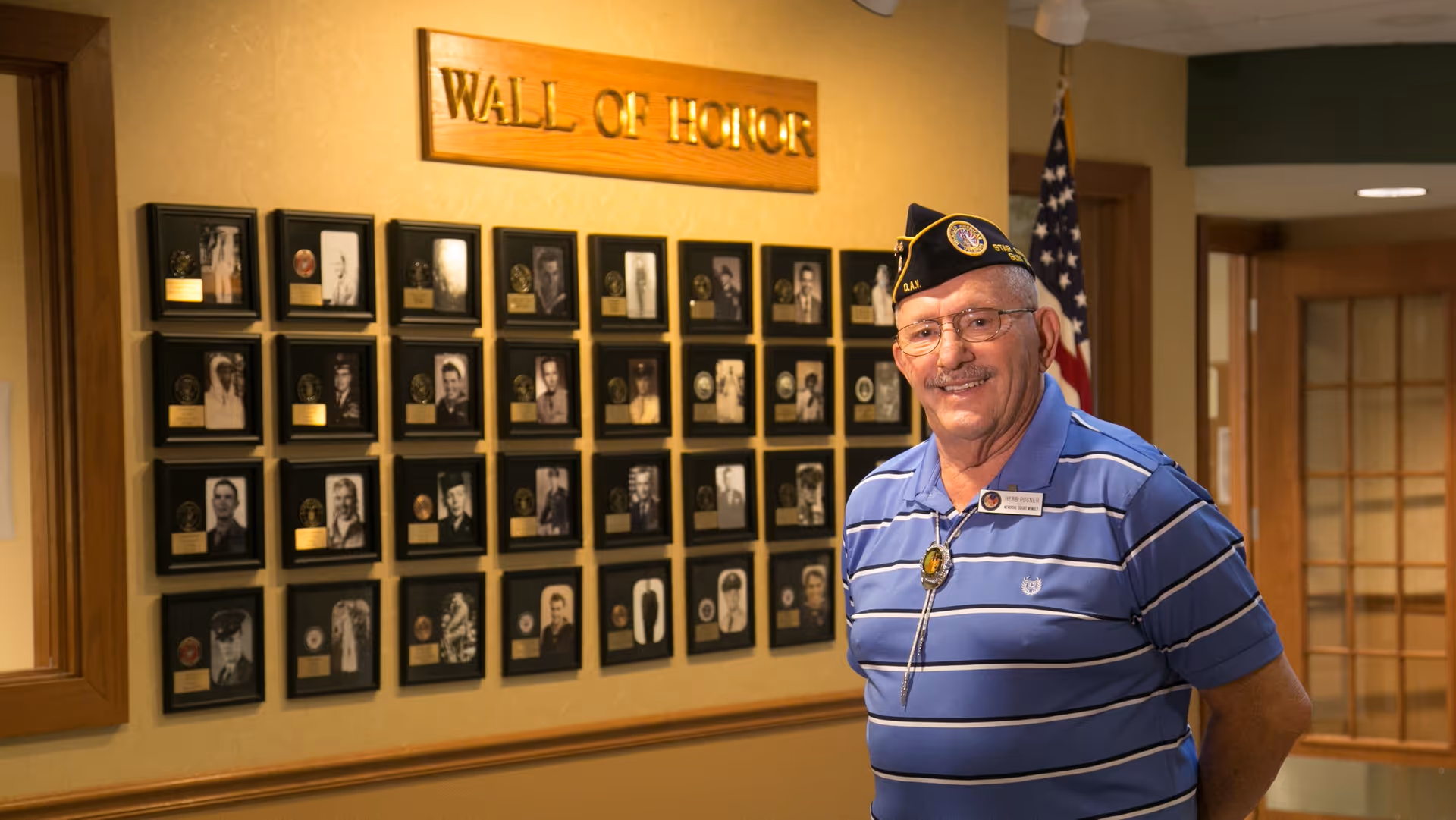 An elderly man wearing a blue striped polo shirt and a veterans cap stands smiling in front of a 'Wall of Honor' display featuring framed photographs and plaques on a beige wall inside a retirement community.
