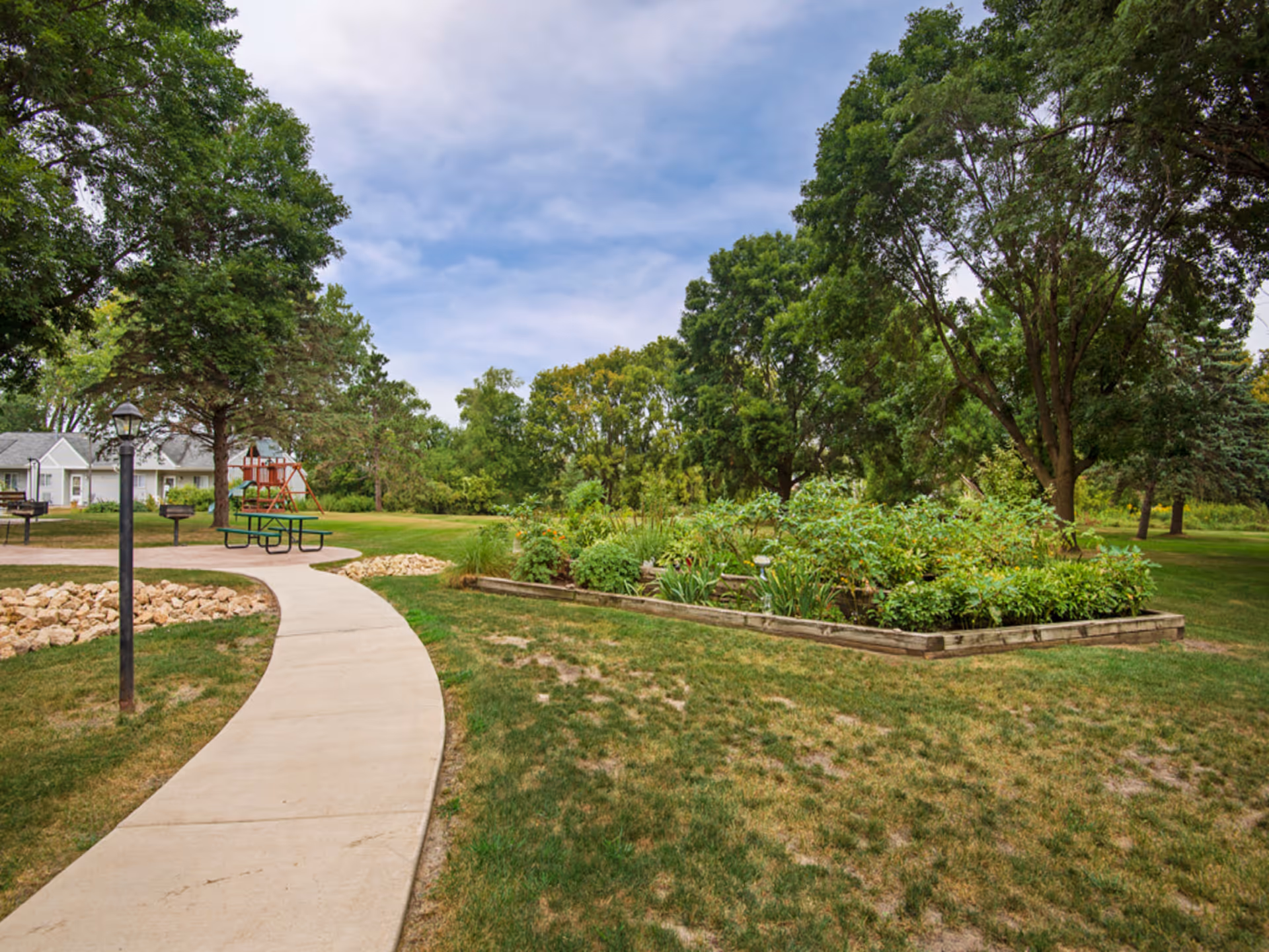 Curved concrete pathway through landscaped outdoor grounds with a picnic table, garden bed, trees, and small residential buildings in the background.