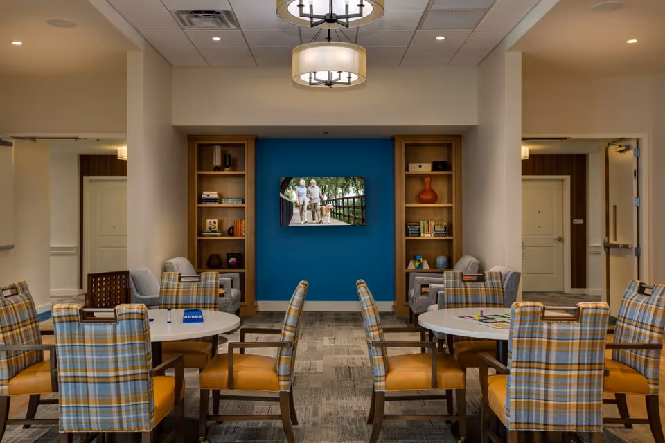 A cozy common area with round tables surrounded by plaid upholstered chairs with yellow cushions. The room features a blue accent wall with a mounted TV displaying an image of two elderly people walking a dog. Wooden bookshelves flank the TV, holding books and decorative items. The space is well-lit with ceiling lights and has a carpeted floor.