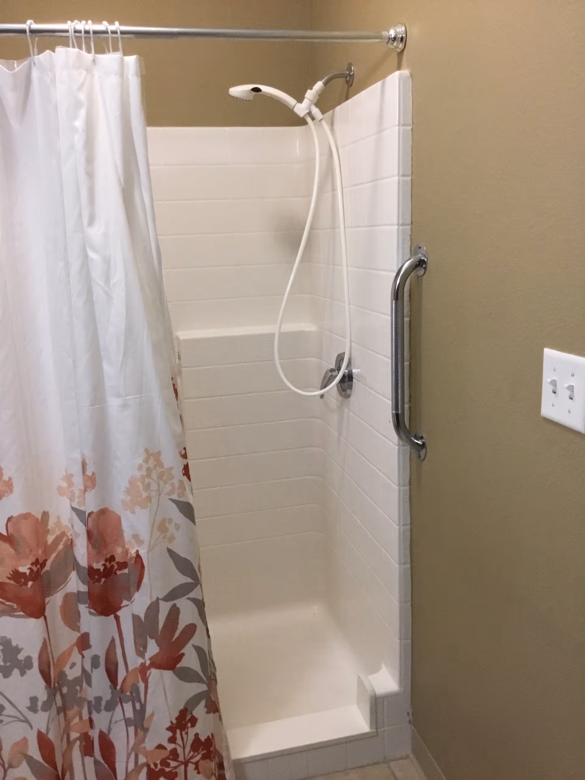 Small tiled shower stall with a handheld showerhead, floral shower curtain, and a wall-mounted grab bar.