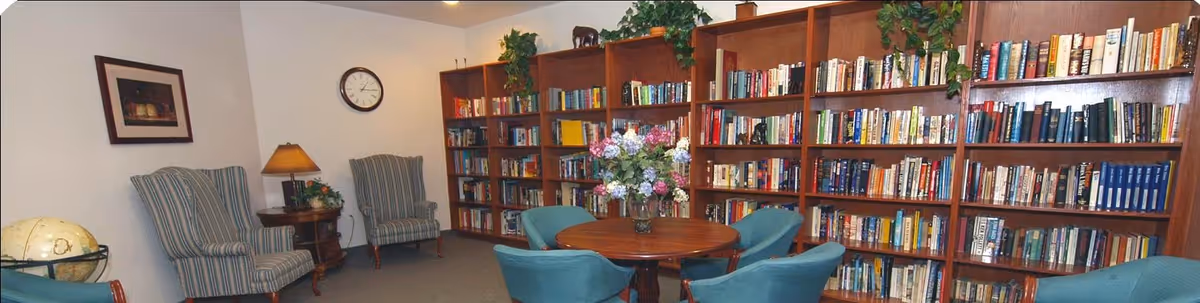 A cozy library room with wooden bookshelves filled with books along the wall, a round wooden table with a vase of flowers in the center, surrounded by blue upholstered chairs. Two striped armchairs and a small side table with a lamp are positioned against the opposite wall, with a clock and framed picture hanging above.