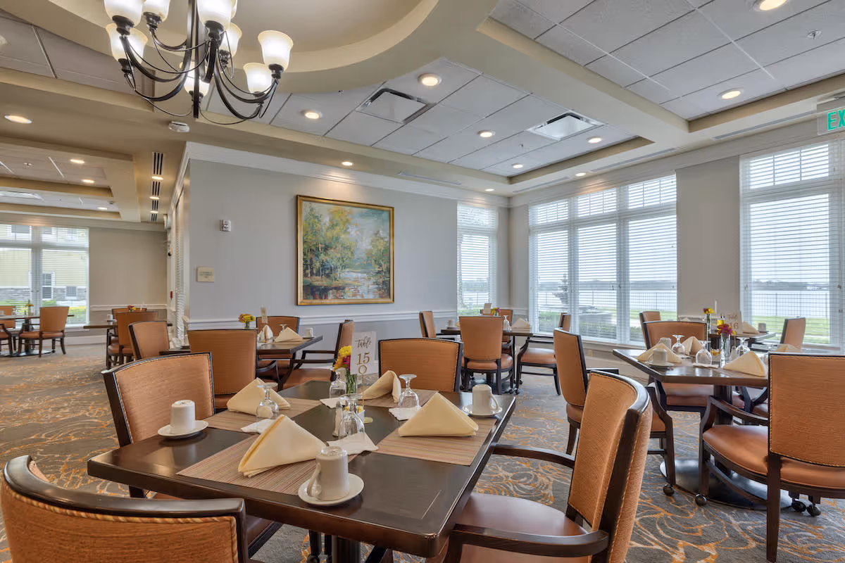 A bright and spacious dining room with multiple tables set with napkins, cups, and glasses. The room features large windows with blinds allowing natural light to fill the space, a chandelier hanging from the ceiling, and a framed painting on the wall. The chairs are cushioned and arranged around dark wooden tables, creating an inviting atmosphere.