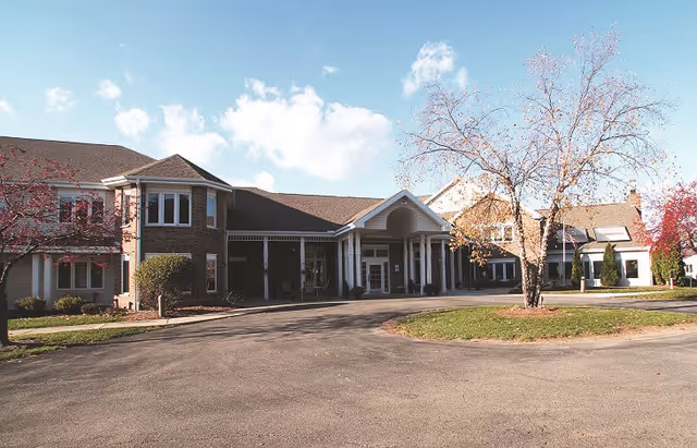 Exterior view of a senior living facility building with a driveway and landscaped area featuring trees with autumn foliage under a partly cloudy sky.