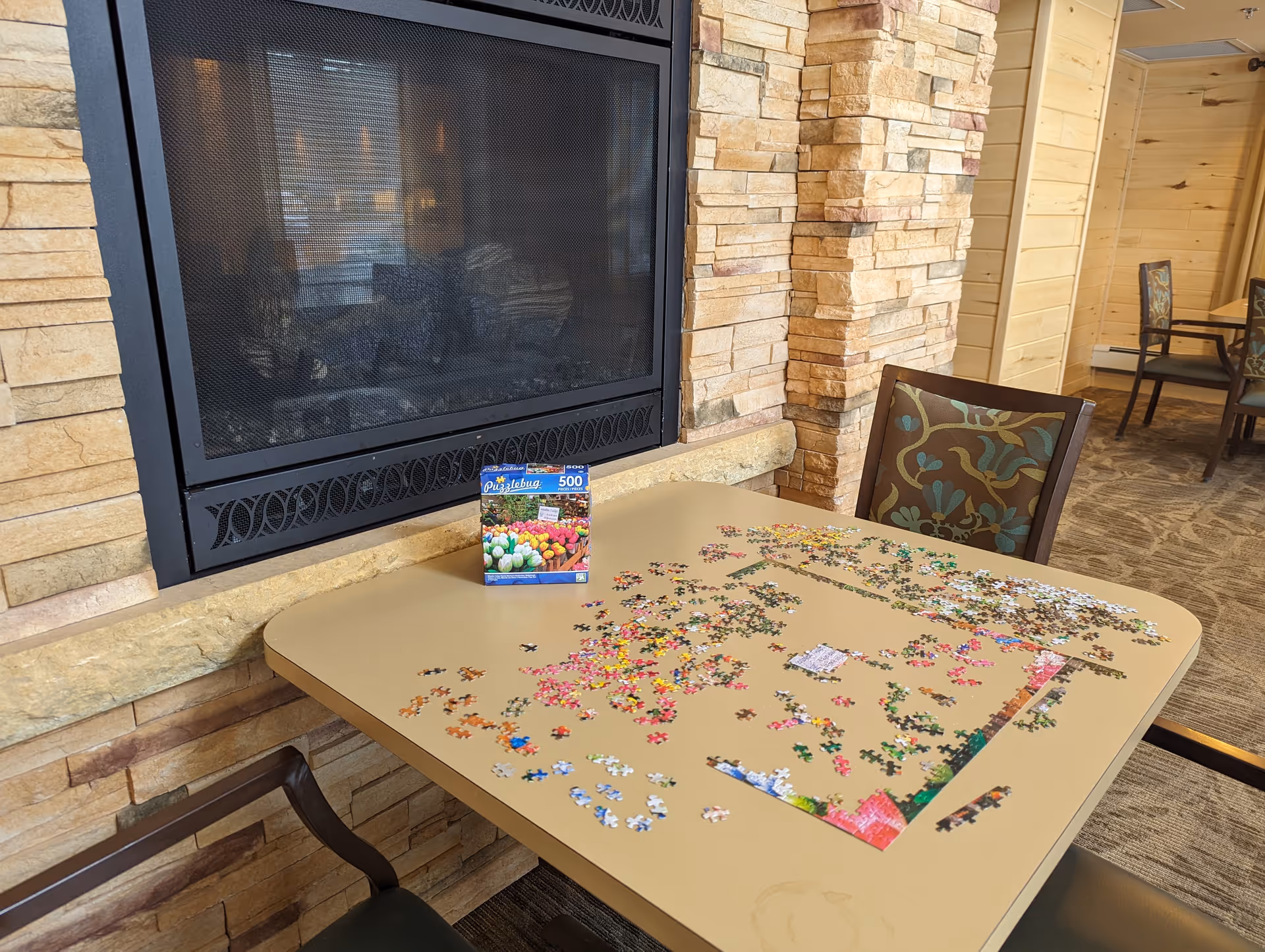 A table with an incomplete 500-piece puzzle featuring colorful tulips, set up in front of a stone fireplace in a cozy room with patterned chairs and wood-paneled walls.