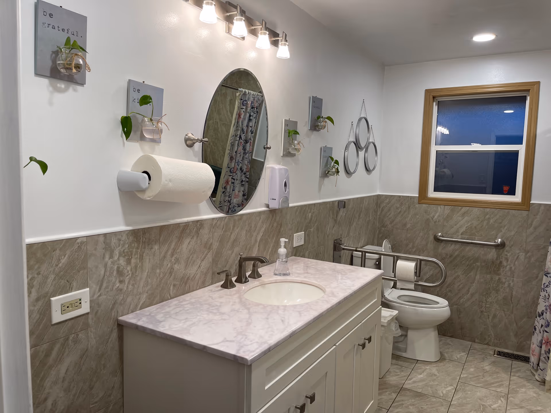 A clean and well-lit bathroom with a white vanity topped with a marble countertop and an oval mirror above it. The walls are half tiled with beige marble tiles and half painted white. There are small wall-mounted planters with green plants and inspirational signs that say 'be grateful.' The bathroom includes a toilet with safety grab bars, a paper towel holder, a soap dispenser, and a window with a wooden frame. A floral shower curtain is partially visible on the right side.