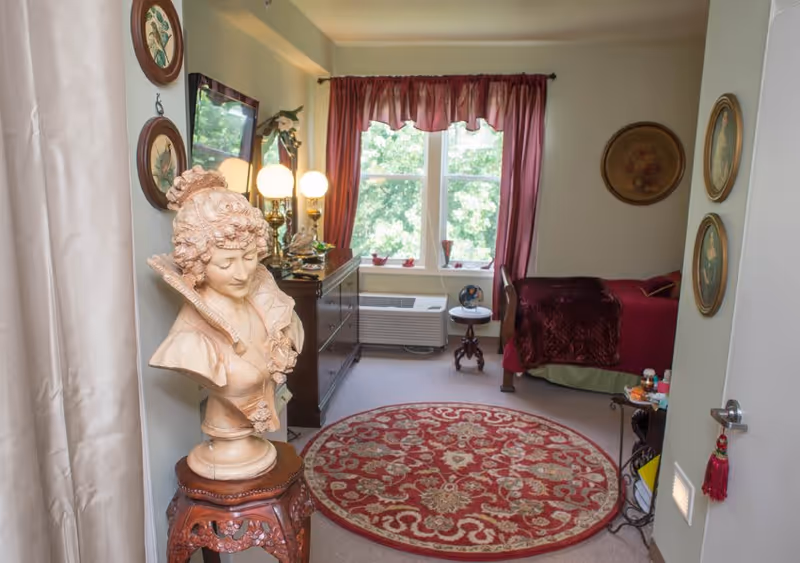 A cozy bedroom with a single bed covered in a dark red blanket, a wooden dresser with a mirror and lamps, a round red patterned rug on the floor, and a decorative bust sculpture on a carved wooden stand near the entrance. The room has two windows with red curtains letting in natural light, and framed artwork on the walls.