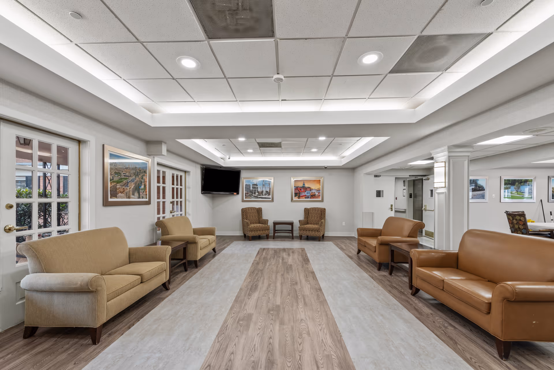 A spacious and well-lit common area in a senior living facility with beige and brown sofas and armchairs arranged along the walls. The room features a modern ceiling with recessed lighting, framed artwork on the walls, a flat-screen TV mounted in the corner, and large windows and glass doors allowing natural light to enter.