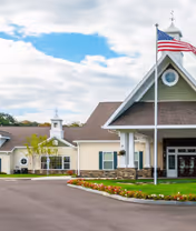 Exterior view of a senior living facility building with a peaked roof, an American flag on a flagpole in front, and a landscaped area with flowers along the driveway under a partly cloudy sky.