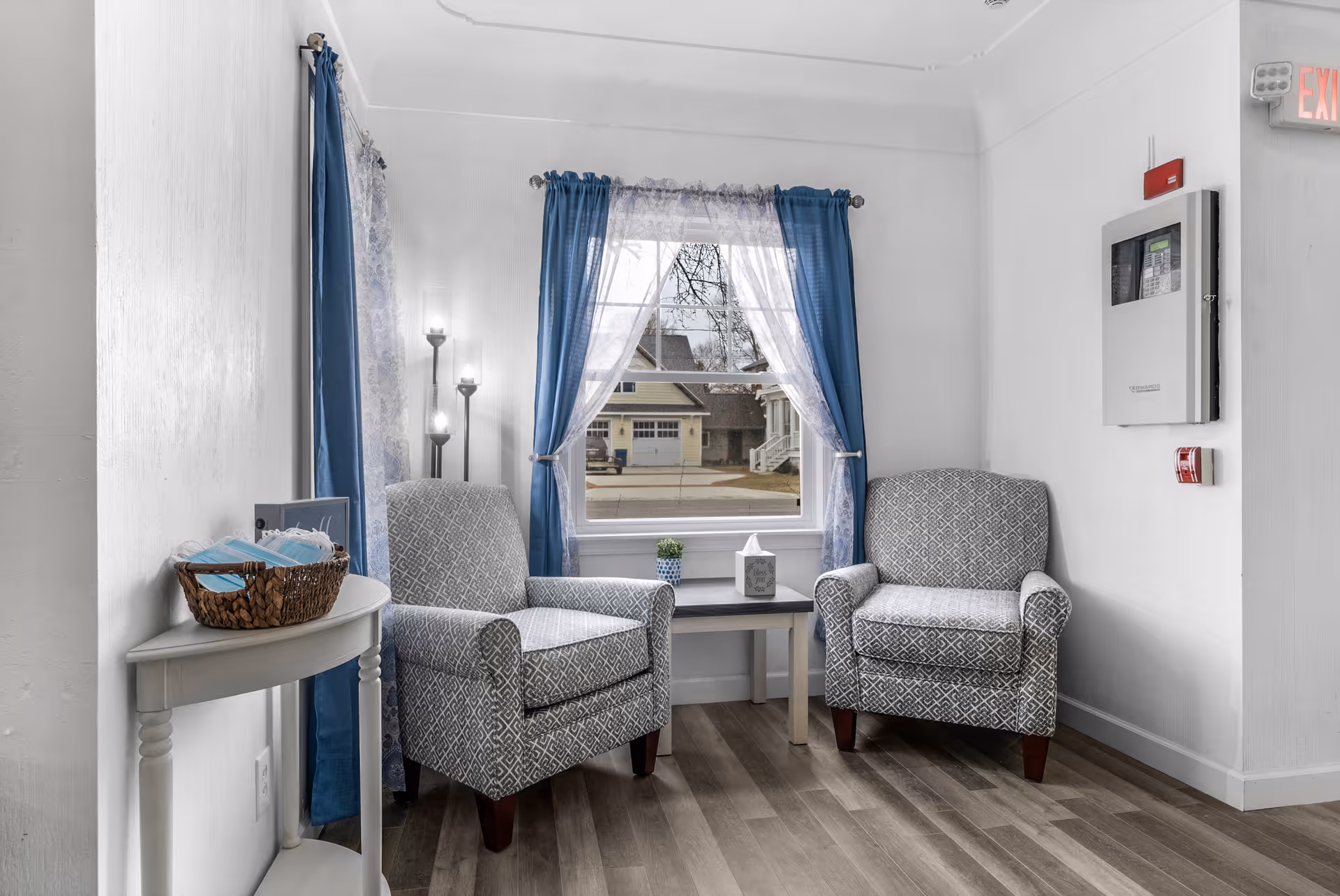 A small sitting area with two patterned armchairs facing each other, separated by a small table with a tissue box and a small plant. The area has a window with blue curtains and sheer white drapes, allowing natural light to brighten the space. A basket with face masks sits on a small white side table to the left. The floor is wood, and there is an emergency exit sign and fire alarm on the wall to the right.