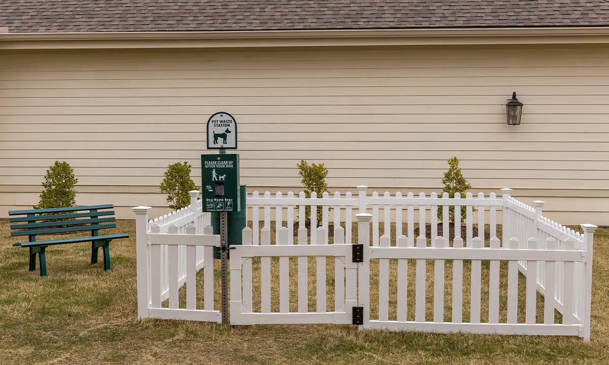 Small white picket fenced pet area with a pet waste station and a green bench in front of a beige-sided building.