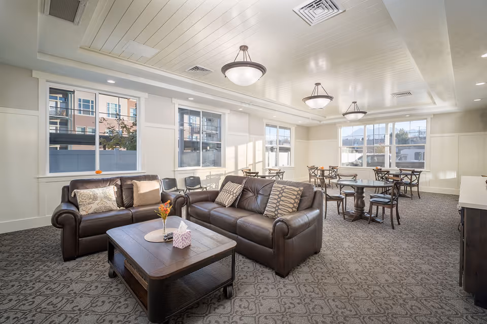 Bright communal lounge with leather sofas, a coffee table, and dining tables by large windows.