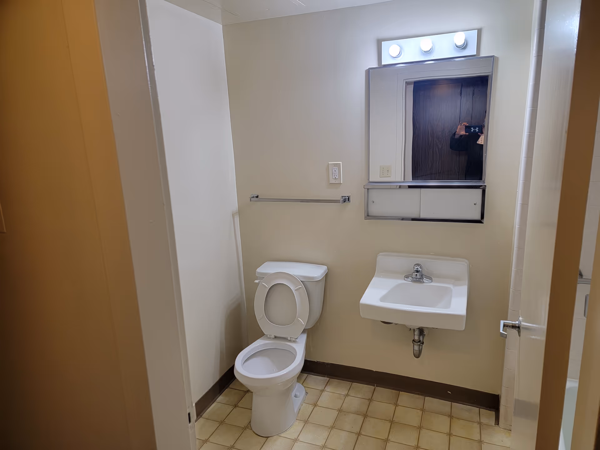 Small bathroom with a toilet, wall-mounted sink, mirror with lights, and tiled floor.