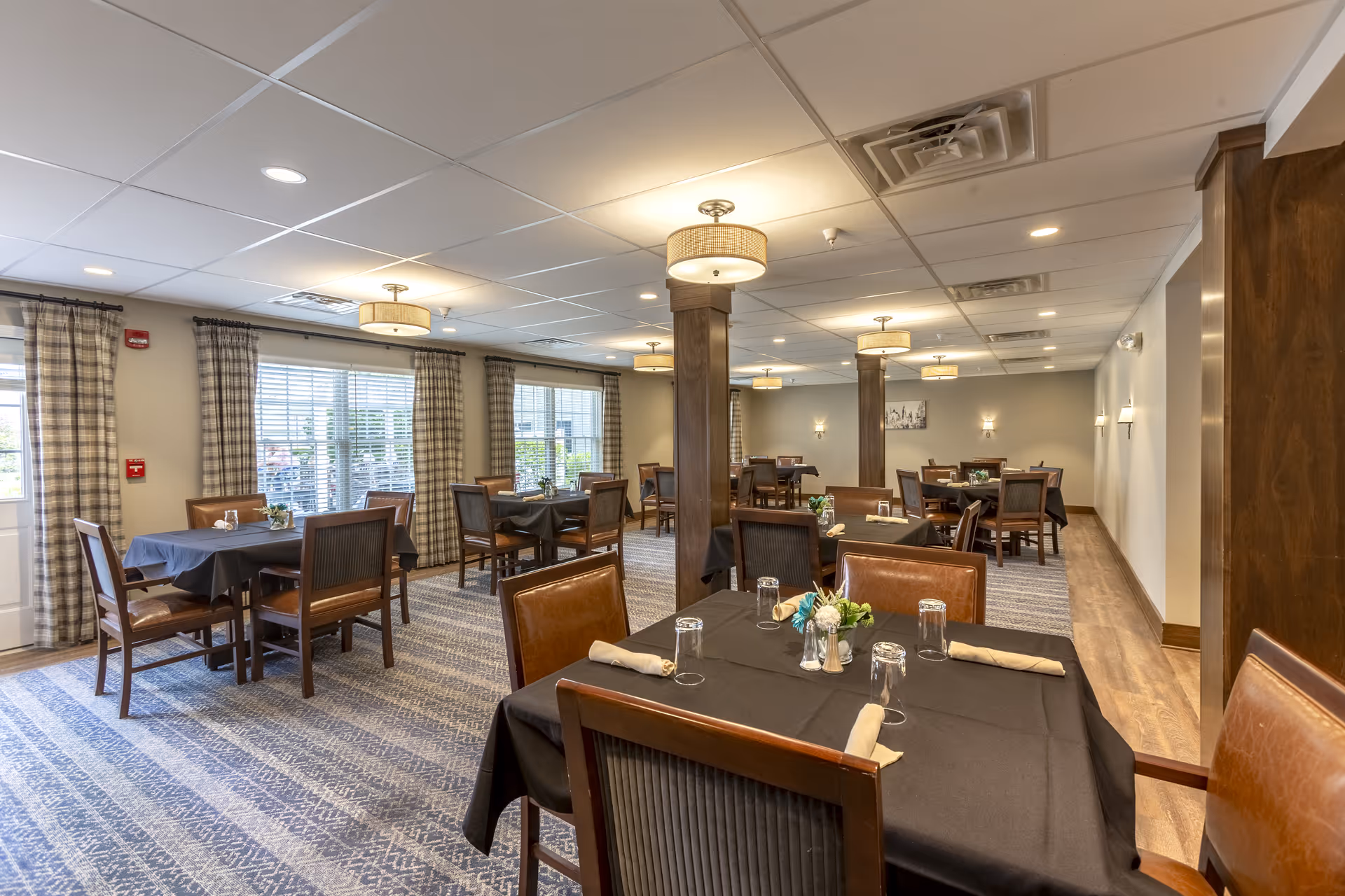 A spacious dining room with multiple tables covered in black tablecloths, each set with glasses, napkins, and small flower arrangements. The room has large windows with plaid curtains, wooden chairs, carpeted floor, and ceiling lights with drum shades.
