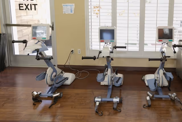 Three stationary exercise bikes with digital screens are positioned on a wooden floor in a room with large windows covered by white blinds. A glass door with an exit sign is visible on the left side.