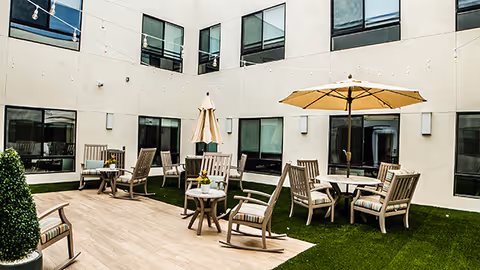 Outdoor courtyard area with wooden chairs and tables, some with umbrellas, surrounded by a multi-story building with large windows. The ground is a combination of artificial grass and wooden decking.