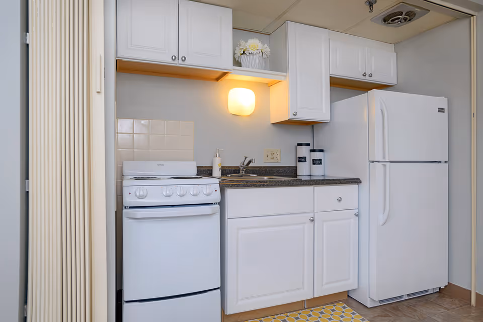 Small kitchen area with white cabinets, a white stove, a white refrigerator, a sink, and a countertop. There is a wall-mounted light fixture above the sink, a small vase with white flowers on the upper shelf, and two labeled canisters on the counter. The floor has a patterned yellow and white rug.