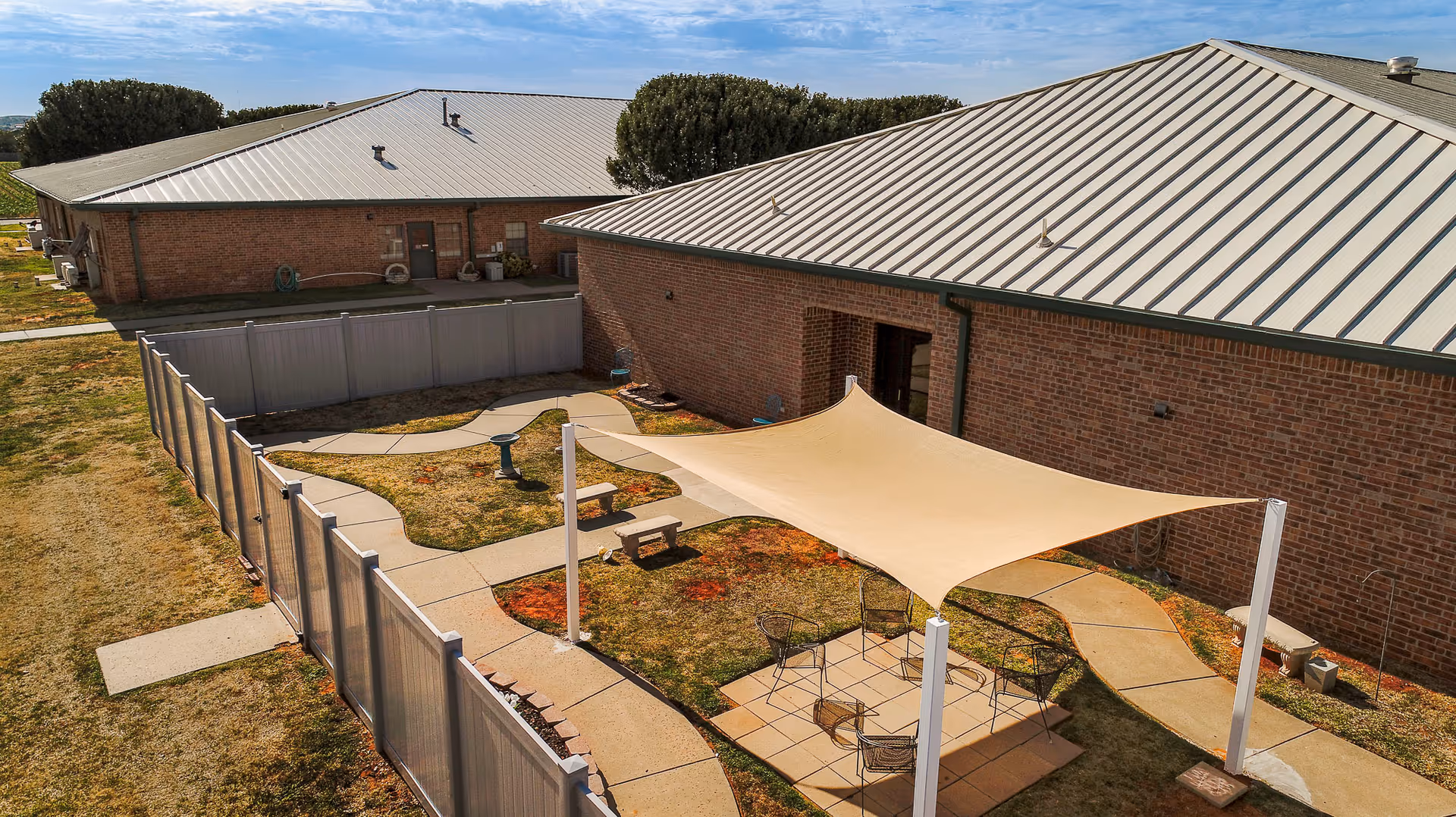 Outdoor fenced patio area at a senior living facility with a beige shade sail canopy, metal chairs, a small table, and a concrete pathway surrounded by grass and two brick buildings with metal roofs.