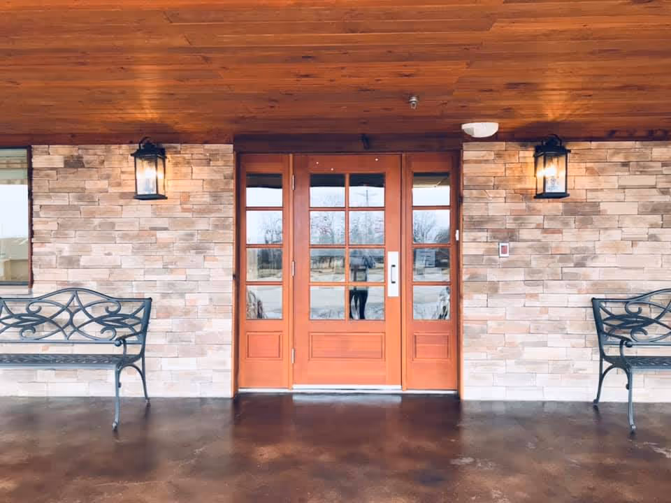 Front entrance of a building with a wooden ceiling, stone brick walls, and a set of double wooden doors with glass panes. Two black metal benches are placed symmetrically on either side of the entrance, and two wall-mounted lantern-style lights are illuminated above the benches.