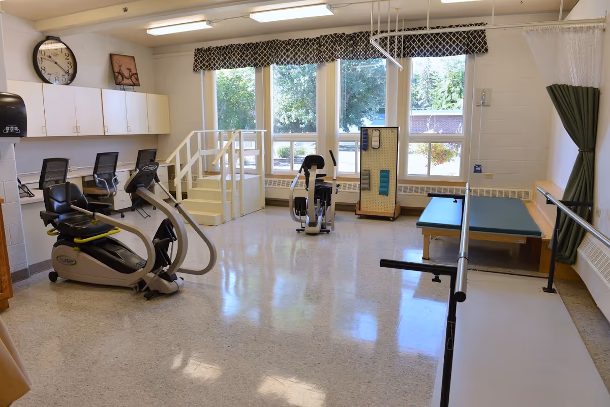 A bright physical therapy or rehabilitation room with exercise equipment including a recumbent bike, handrails for walking practice, a padded therapy table, and a small set of stairs. Large windows with patterned valances let in natural light, and there are chairs and cabinets along one wall.