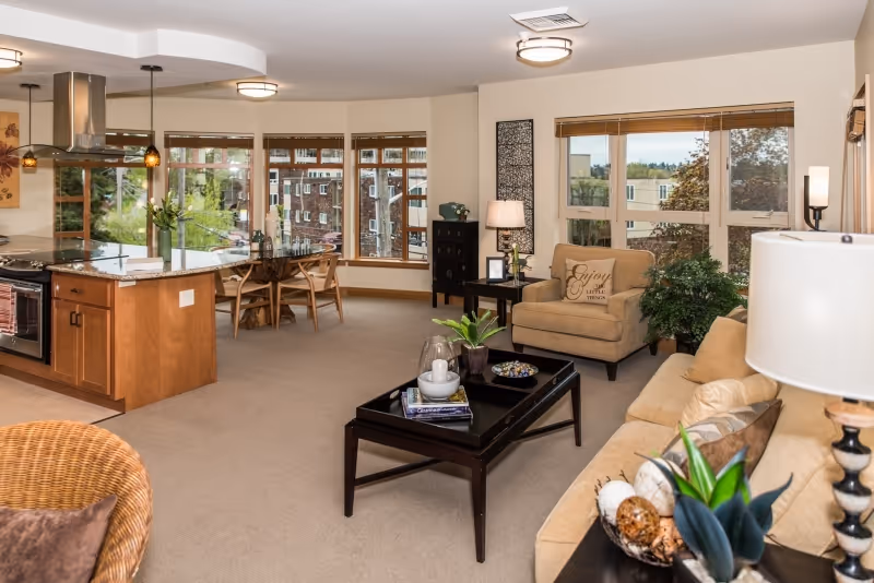 Open-concept living area with a sofa, armchair, coffee table, and a kitchen island with a dining table by large windows.